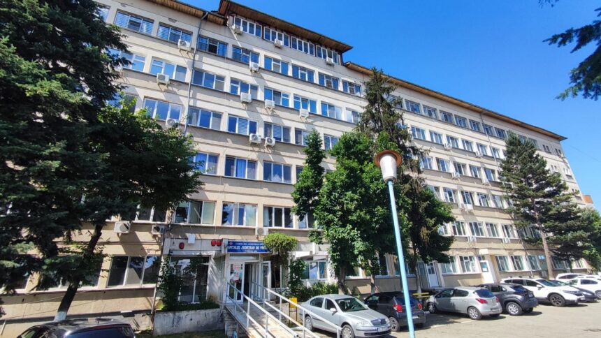 Facade of a large beige, multi-story hospital/official building with many windows, entrance with a ramp, parked cars, and surrounding trees under a clear blue sky.