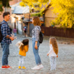 Man and woman with two young daughters converse on a sunlit cobblestone street; kids hold plush toys, autumn trees in background.