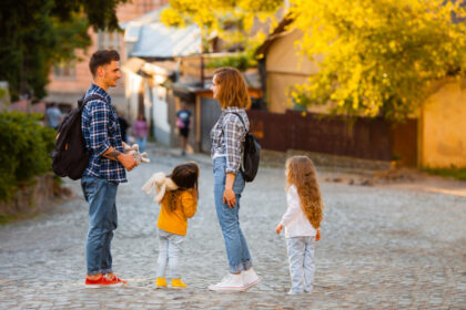 Man and woman with two young daughters converse on a sunlit cobblestone street; kids hold plush toys, autumn trees in background.
