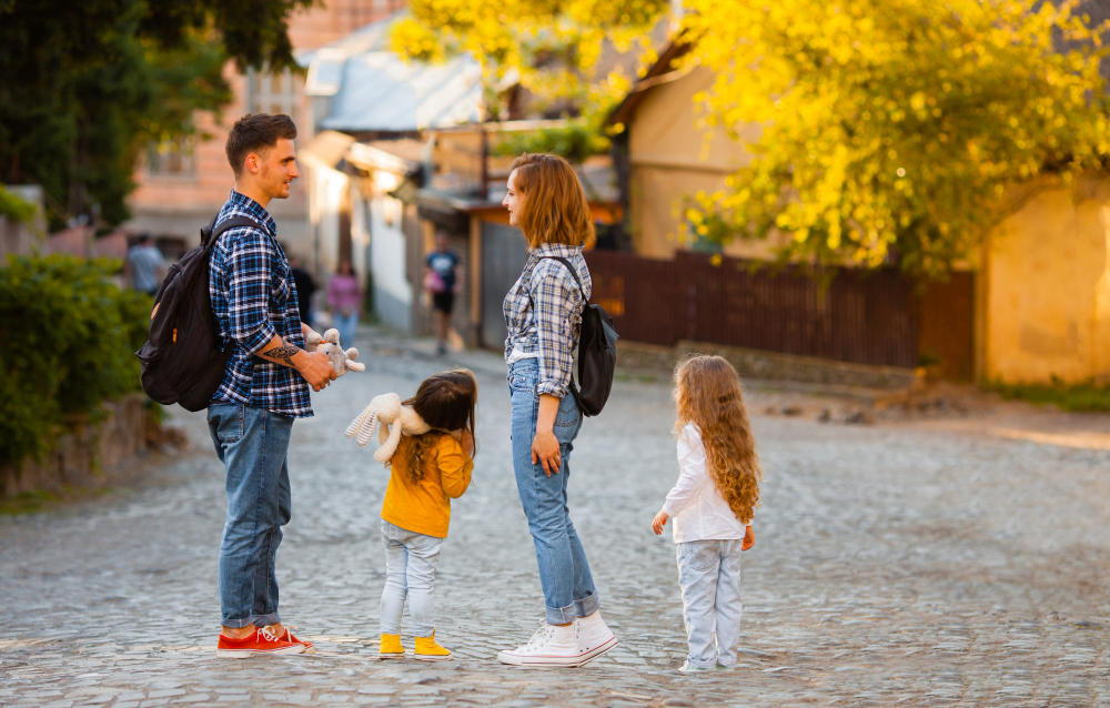Man and woman with two young daughters converse on a sunlit cobblestone street; kids hold plush toys, autumn trees in background.