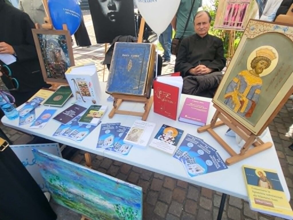Outdoor book booth with a table full of pamphlets and books, framed artwork, and a man seated in the background.