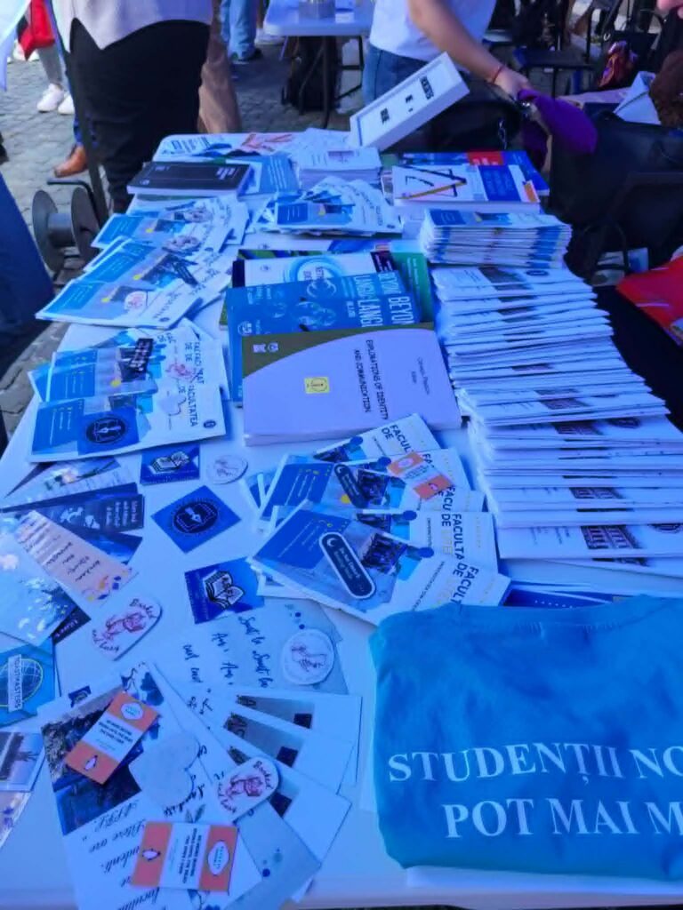 Table spread with blue pamphlets and booklets at an outdoor event; a blue tote bag on the right and people in the background.