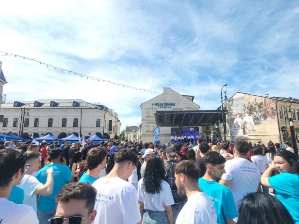 Large crowd of people wearing blue/white shirts gathers in a city square with a stage in the background.
