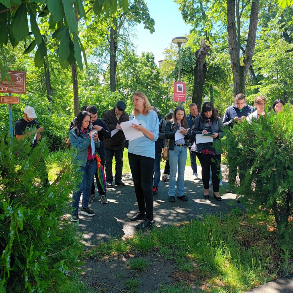 Group of people standing on a park path, reading and signing papers among green bushes and trees on a sunny day.