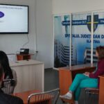 Presenter points to a slide on a wall-mounted screen during a hospital training seminar, attendees seated in front of her.