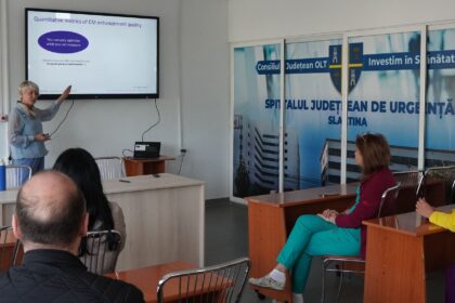 Presenter points to a slide on a wall-mounted screen during a hospital training seminar, attendees seated in front of her.