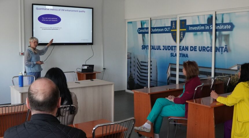 Presenter points to a slide on a wall-mounted screen during a hospital training seminar, attendees seated in front of her.