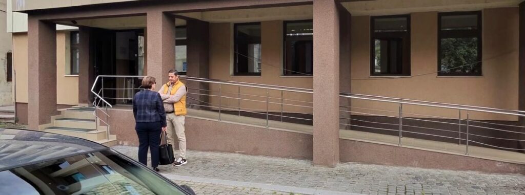 Two adults chat outside a tan building with a ramped entrance and a parked car nearby.
