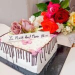 White square birthday cake with chocolate drip and pink flower decorations, with a colorful rose bouquet in the background behind it.