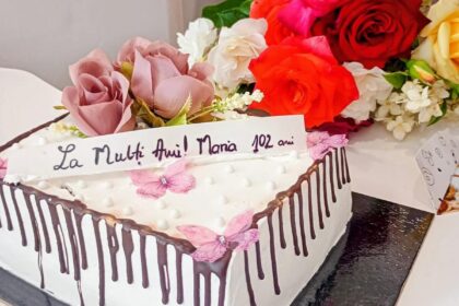 White square birthday cake with chocolate drip and pink flower decorations, with a colorful rose bouquet in the background behind it.