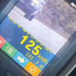 Car dashboard screen showing current speed 125 km/h and distance 181.3 m on a blue/orange display with a truck ahead on a road.