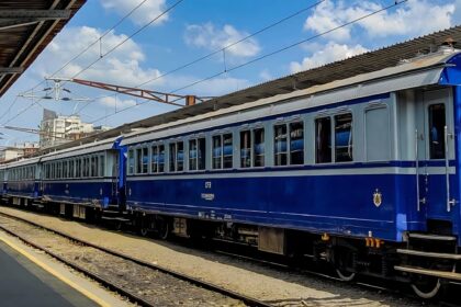 Blue passenger train parked at a station beside the platform under a clear, sunny sky with overhead power lines above.