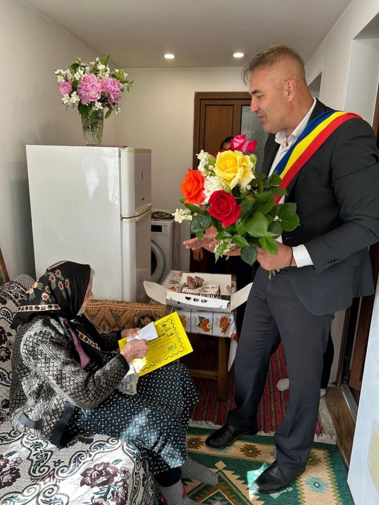 A man in a suit with a ceremonial sash hands a colorful bouquet to an elderly woman seated on a sofa in a cozy living room.