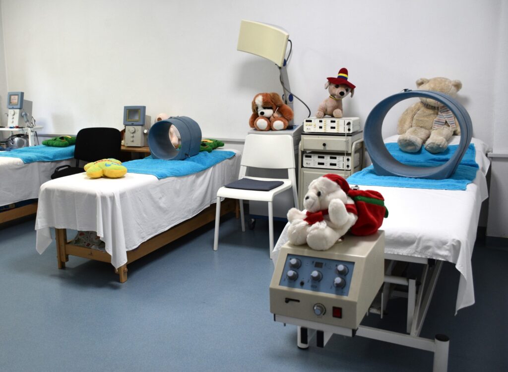 Playful children’s hospital scene with two beds, blue towels, and stuffed animals near medical equipment on the wall and carts.