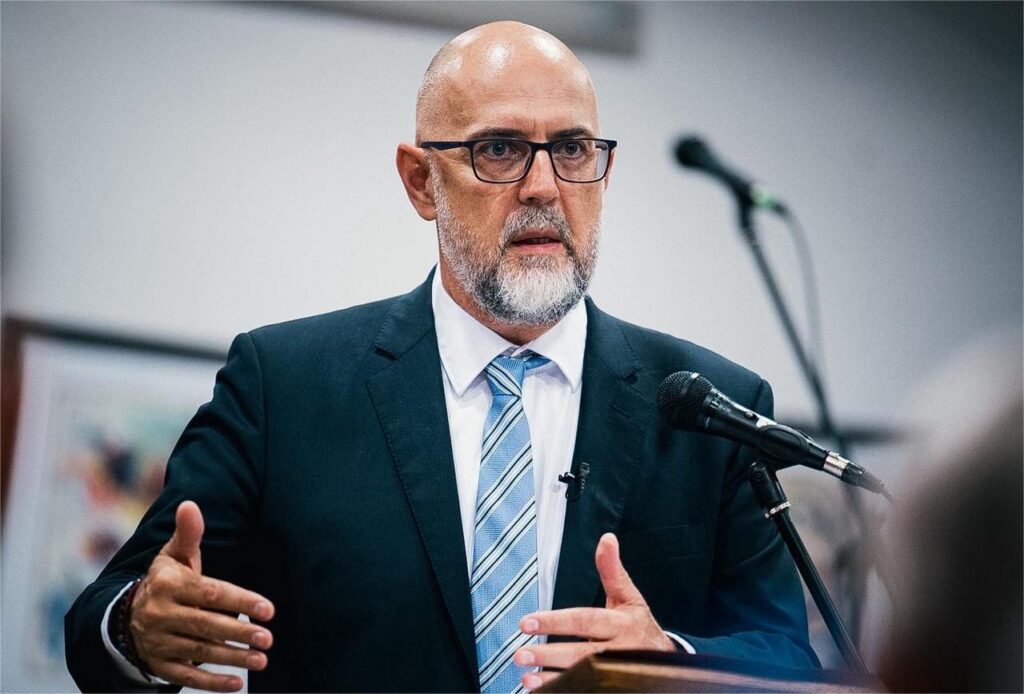 Kelemen Hunor. Foto: Facebook - Jurnalul Olteniei – Știri din Craiova și Oltenia Bald man with glasses and beard in a dark suit speaking at a podium with a microphone.