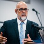 Bald man with glasses and beard in a dark suit speaking at a podium with a microphone.