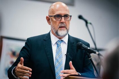 Bald man with glasses and beard in a dark suit speaking at a podium with a microphone.