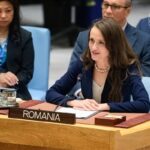 Romanian delegate woman speaks into a desk microphone at a United Nations–style meeting, Romania nameplate in foreground.