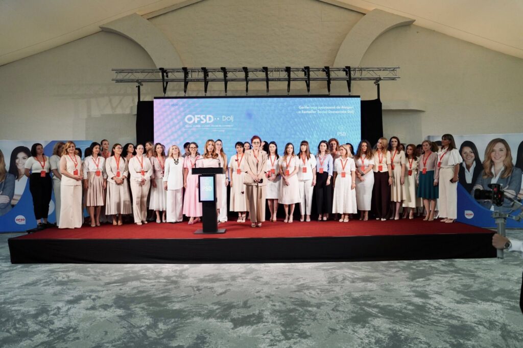 Rareș Bogdan, mesaj neașteptat pentru OFSD Dolj. Foto: Facebook - Jurnalul Olteniei – Știri din Craiova și Oltenia Group of women in light clothing standing on a stage with a blue backdrop and red carpet, posing for a conference photo.
