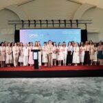 Group of women in light clothing standing on a stage with a blue backdrop and red carpet, posing for a conference photo.
