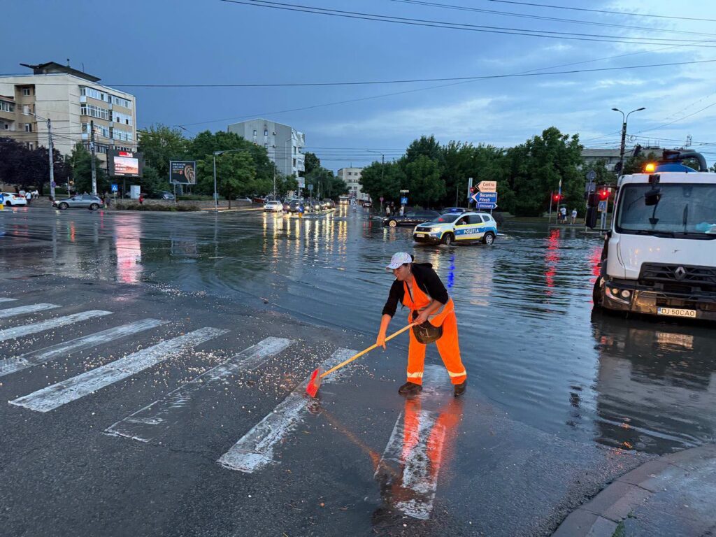 City street heavily flooded; a worker in orange reflective gear sweeps water from the crosswalk with a broom while a police car and trucks are in the background.