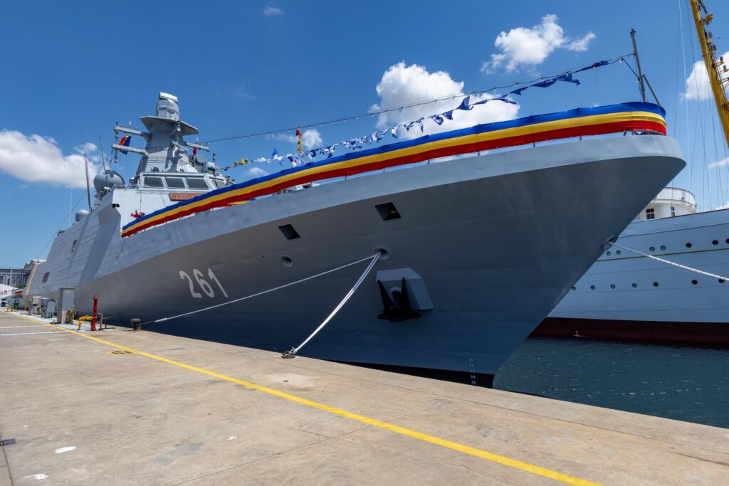 - Jurnalul Olteniei – Știri din Craiova și Oltenia Large gray naval ship moored at a pier, bow decorated with blue, yellow, and red stripe under a blue sky and white clouds.