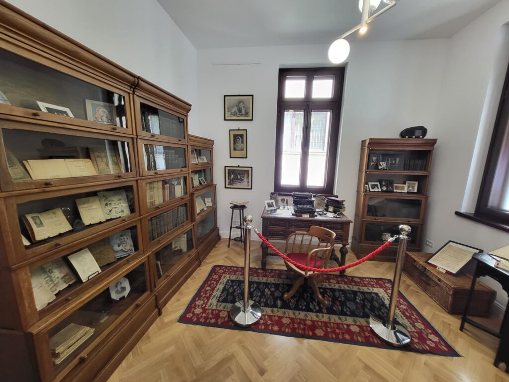 Casa Memorială Elena Farago - Jurnalul Olteniei – Știri din Craiova și Oltenia Vintage study with glass-front wooden bookcases, a desk with a typewriter, and framed photos by a window in a museum-like room.
