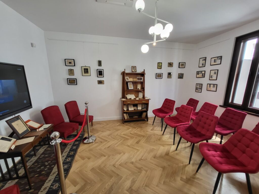 Casa Memorială Elena Farago a fost reabilitată - Jurnalul Olteniei – Știri din Craiova și Oltenia Reading room with red tufted chairs arranged in a semi-circle, a wooden bookcase displaying documents, and framed pictures on white walls.