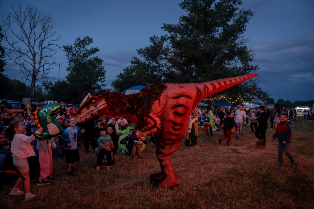 Crowd of children and adults at an outdoor event watching a large red dinosaur puppet perform at dusk.