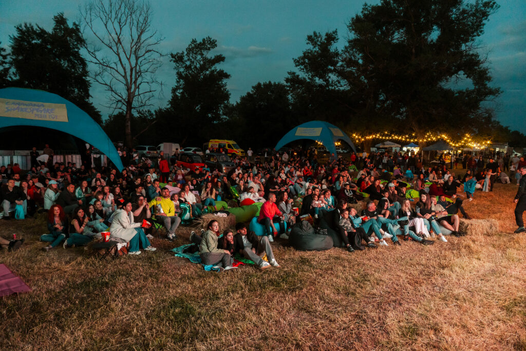 Large crowd seated on the grass at an outdoor festival, with tents, string lights, and trees in the background.