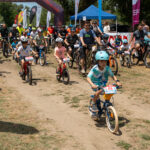Group of children and adults riding bicycles at a race start on a dirt path, with banners and a blue tent in the background.