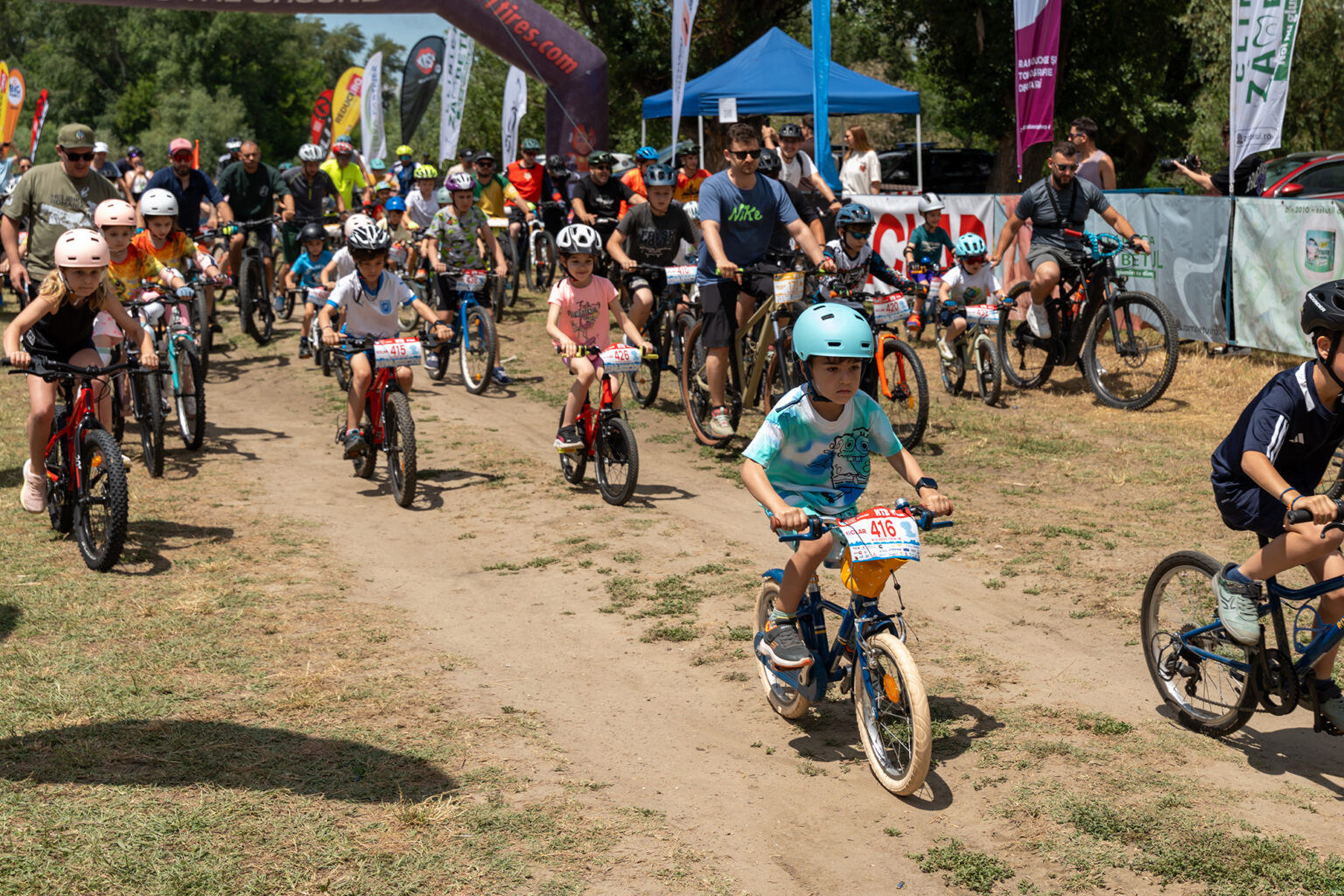 Group of children and adults riding bicycles at a race start on a dirt path, with banners and a blue tent in the background.