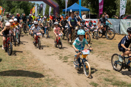 Group of children and adults riding bicycles at a race start on a dirt path, with banners and a blue tent in the background.