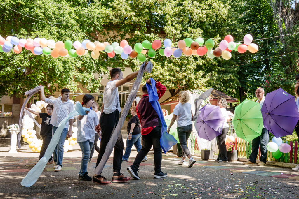 FOTO-115 - Jurnalul Olteniei – Știri din Craiova și Oltenia People dancing and waving colorful fabric ribbons under a string of pastel balloons at an outdoor celebration.