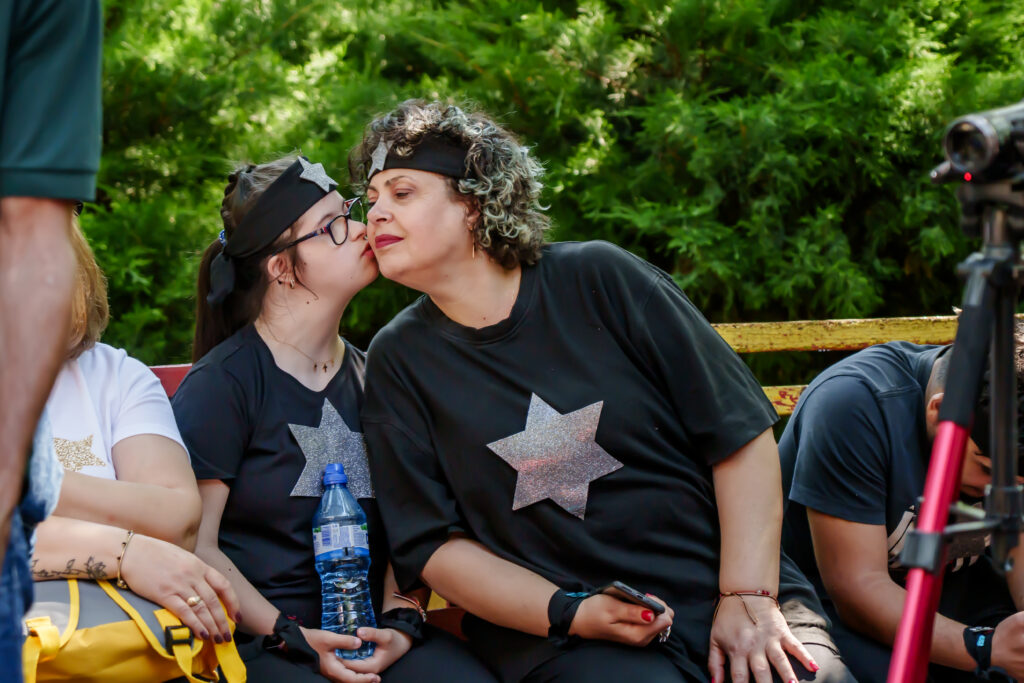 FOTO-159 - Jurnalul Olteniei – Știri din Craiova și Oltenia Two women in matching black shirts with glitter stars sit on a bench outdoors; one kisses the other on the cheek, showing a warm moment of friendship.