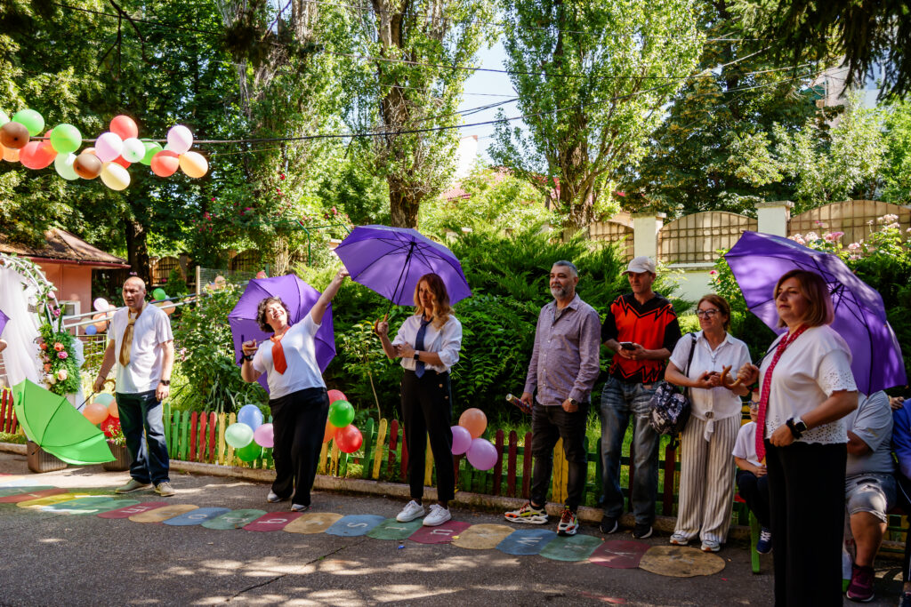 FOTO-34 - Jurnalul Olteniei – Știri din Craiova și Oltenia Group of adults in a sunny garden holding purple umbrellas during a festive outdoor party with balloons and colorful ground decorations.