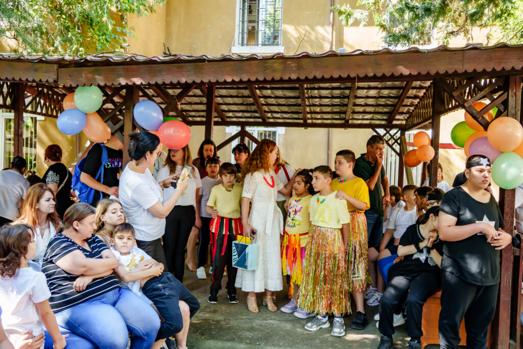 FOTO-49 - Jurnalul Olteniei – Știri din Craiova și Oltenia Crowd of children and adults at an outdoor celebration under a wooden gazebo, balloons decorating the space.