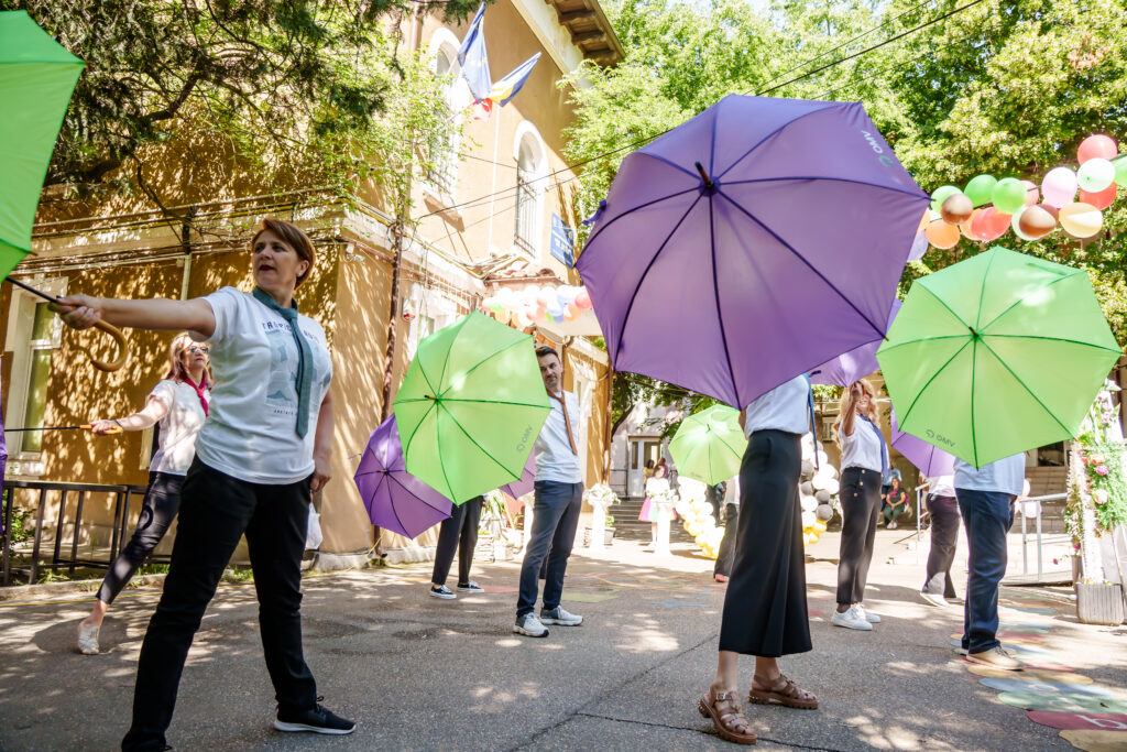 FOTO-71 - Jurnalul Olteniei – Știri din Craiova și Oltenia Parade performers in white shirts holding large green and purple umbrellas on a sunny street, with balloons overhead beside a beige building.