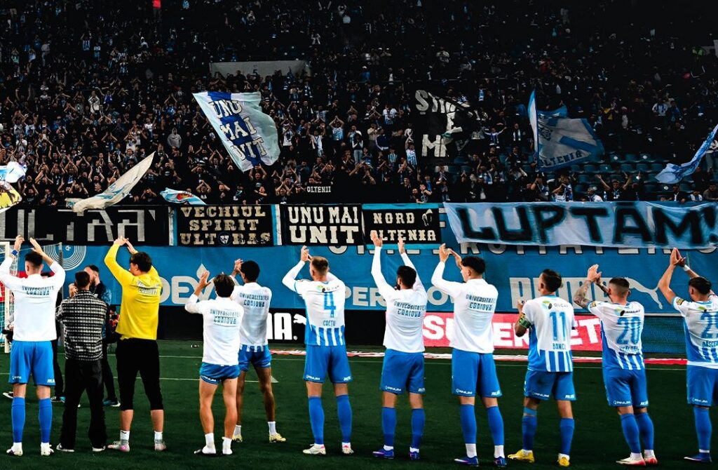 LPF a ales cei mai valoroși tineri din Superliga României Universitatea Craiova se laudă cu 2 jucători în formația U21 - Jurnalul Olteniei – Știri din Craiova și Oltenia Soccer team in white and blue uniforms stands in a line on the field as fans cheer with banners and flags in the stands.