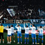 Soccer team in white and blue uniforms stands in a line on the field as fans cheer with banners and flags in the stands.