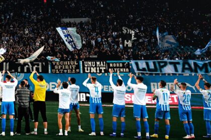 Soccer team in white and blue uniforms stands in a line on the field as fans cheer with banners and flags in the stands.