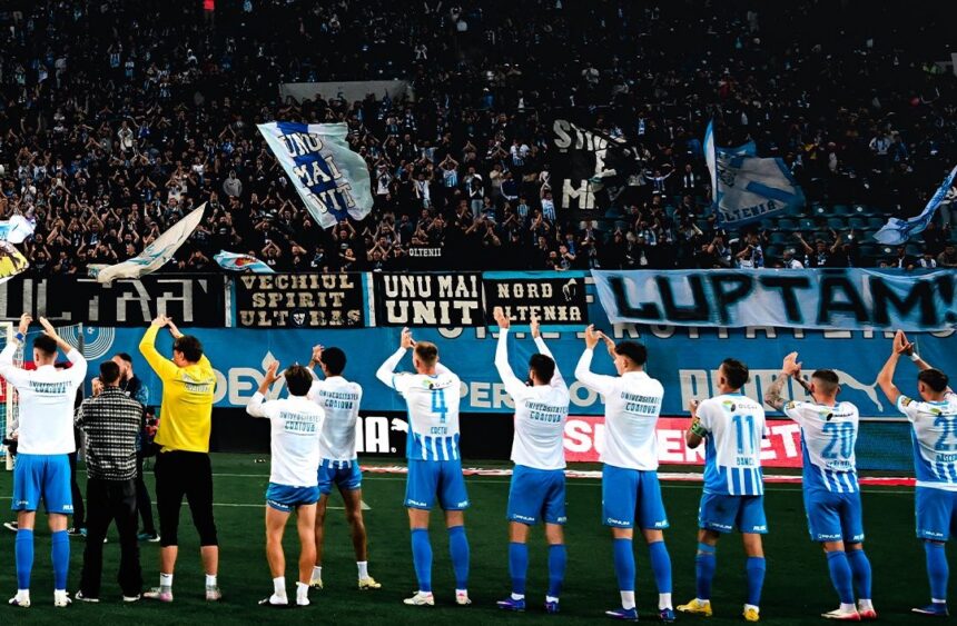 Soccer team in white and blue uniforms stands in a line on the field as fans cheer with banners and flags in the stands.