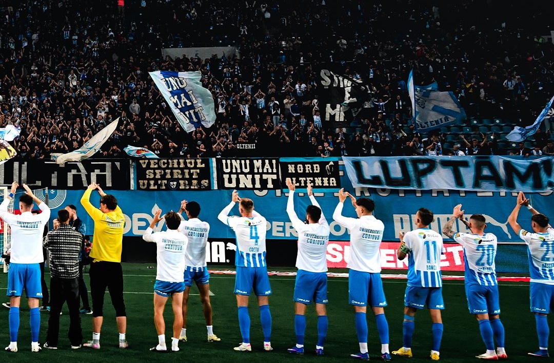Soccer team in white and blue uniforms stands in a line on the field as fans cheer with banners and flags in the stands.