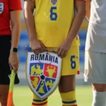 Three soccer players standing side by side: red referee shirt, yellow Romania kit, and gray kit, with the central player holding a shield badge reading ROMANIA.