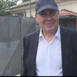Man in a dark suit and white shirt wearing a blue cap, standing outside a government building with a round Ministerul Justiției seal on a gray metal gate in the background.
