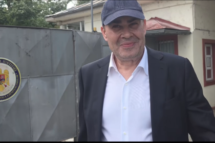 Man in a dark suit and white shirt wearing a blue cap, standing outside a government building with a round Ministerul Justiției seal on a gray metal gate in the background.