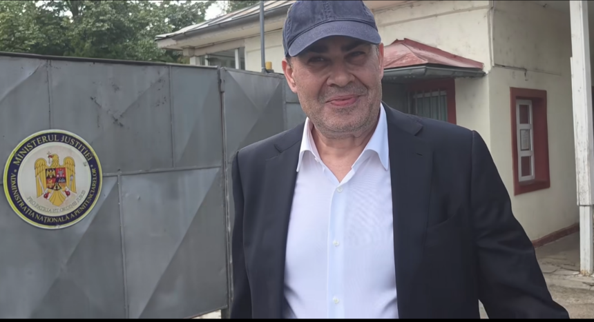 Man in a dark suit and white shirt wearing a blue cap, standing outside a government building with a round Ministerul Justiției seal on a gray metal gate in the background.