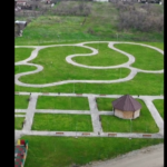 Aerial view of a park with winding white gravel paths around a small hexagonal gazebo and neat grassy plots with benches.