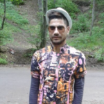 Man wearing a colorful patterned shirt, sunglasses, and a light hat standing on a forest road.