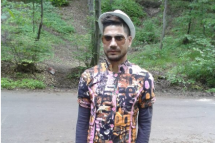 Man wearing a colorful patterned shirt, sunglasses, and a light hat standing on a forest road.
